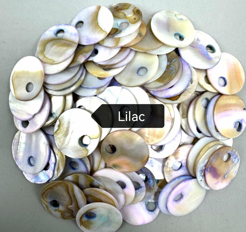 May include: A collection of round, flat shell discs with iridescent surfaces in shades of white, cream, tan, and soft purple. Each disc features a single circular hole near the edge. A black rectangular label with the white text Lilac is centered.