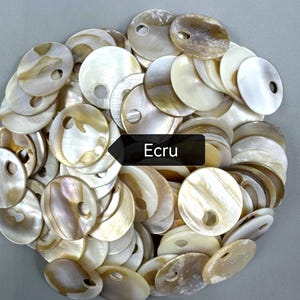 May include: A collection of round, flat mother-of-pearl buttons in shades of cream, beige, and off-white. Each button features a single circular hole near the edge. A black rectangular label with the white text Ecru is centered on the pile.