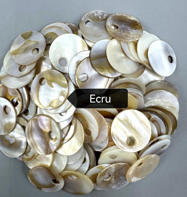 May include: A collection of round, flat mother-of-pearl buttons in shades of cream, beige, and off-white. Each button features a single circular hole near the edge. A black rectangular label with the white text Ecru is centered on the pile.