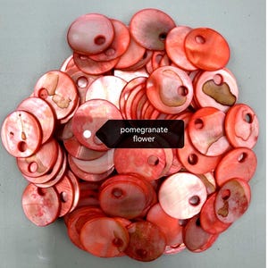 May include: A collection of round, flat, red-toned shell buttons, each featuring a single off-center hole. The surfaces exhibit a natural, iridescent sheen with varied organic textures and markings. A black label displays the text pomegranate flower.