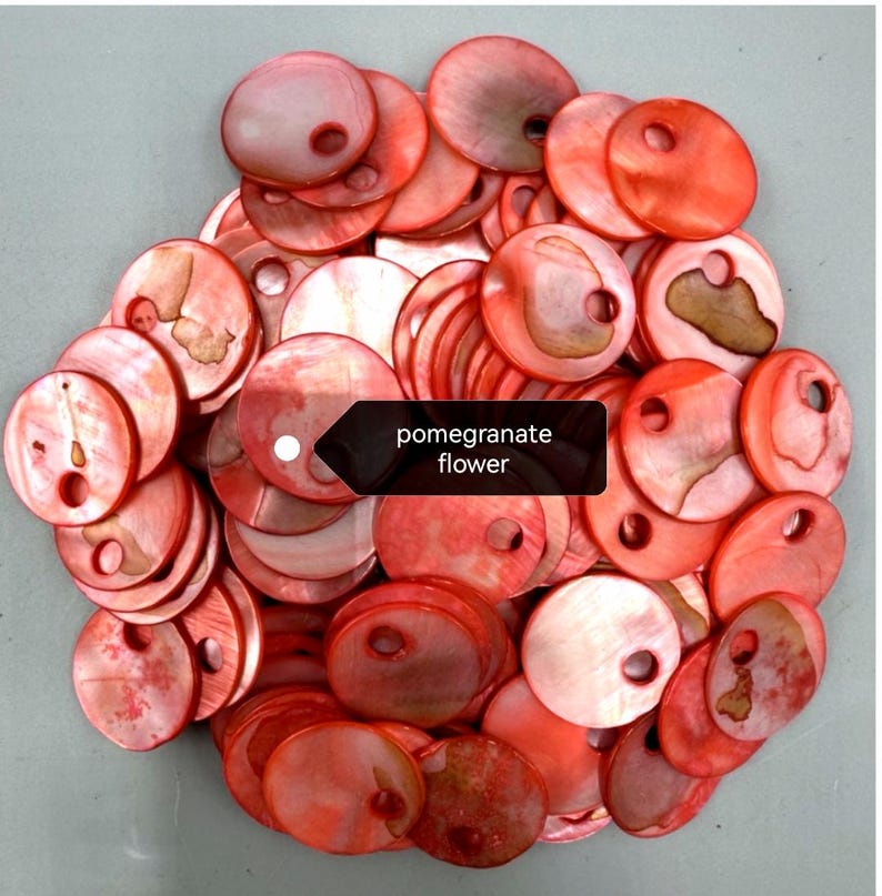 May include: A collection of round, flat, red-toned shell buttons, each featuring a single off-center hole. The surfaces exhibit a natural, iridescent sheen with varied organic textures and markings. A black label displays the text pomegranate flower.