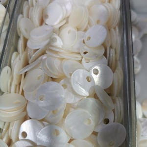 May include: A collection of small, round, off-white mother-of-pearl buttons, each featuring a single circular hole near the edge. These flat, iridescent shell discs are stacked together in a clear container, showcasing their natural luster.