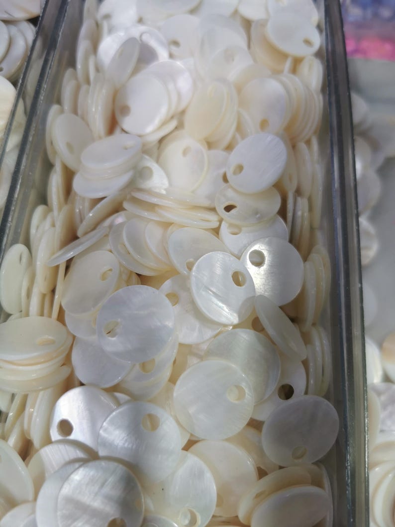 May include: A collection of small, round, off-white mother-of-pearl buttons, each featuring a single circular hole near the edge. These flat, iridescent shell discs are stacked together in a clear container, showcasing their natural luster.