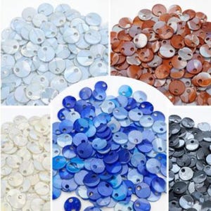 Peut inclure: Une collection de petits disques plats et ronds avec un seul trou près du bord, présentés en cinq groupes de couleurs distincts : blanc, marron, crème, bleu et noir. Chaque disque présente une surface lisse et réfléchissante avec un éclat irisé.