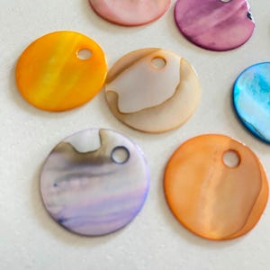 Peut inclure: Une collection de disques en coquillage plats et circulaires de différentes couleurs, notamment orange, jaune, violet, bleu et blanc irisé. Chaque disque présente un petit trou circulaire près du bord supérieur, avec une surface lisse et polie.