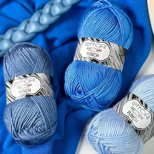 Op de afbeelding: Een assortiment blauwe bollen garen met labels "etrofil PUNCH". Het garen is in verschillende blauwtinten, van licht tot donker, en is gerangschikt op een blauwe stoffen achtergrond. Een blauwe breinaald is ook zichtbaar.