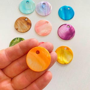 Può includere: Una collezione di ciondoli circolari e piatti in conchiglia iridescente in vari colori, tra cui arancione, giallo, blu, viola, rosa, marrone e verde. Ogni ciondolo presenta un piccolo foro vicino al bordo superiore per l'attacco o l'infilatura.