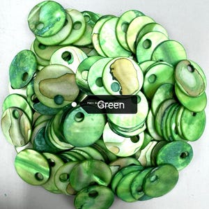 May include: A large collection of circular, flat, green-dyed shell buttons, each featuring a single off-center hole for attachment. The buttons display natural iridescent textures and variations in green tones, with some showing tan patches.