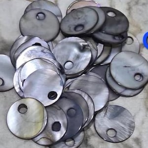 May include: A collection of circular, iridescent gray mother-of-pearl buttons, each featuring a single off-center hole. The buttons display a natural, shimmering finish with varied dark and light gray tones, and the partial blue text GR is visible.