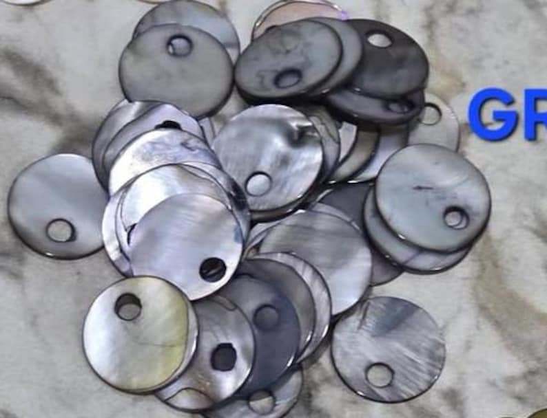 May include: A collection of circular, iridescent gray mother-of-pearl buttons, each featuring a single off-center hole. The buttons display a natural, shimmering finish with varied dark and light gray tones, and the partial blue text GR is visible.