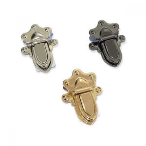 May include: Three metal clasps in silver, black, and gold. Each clasp has a crown-like top and a curved, boat-shaped body. The clasps are designed for use in bags, boxes, or other craft projects.