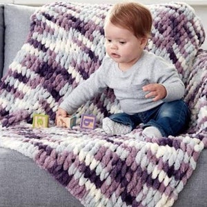 May include: A baby sitting on a couch with a purple and white striped knitted blanket. The blanket is made of chunky yarn and has a soft, fluffy texture.