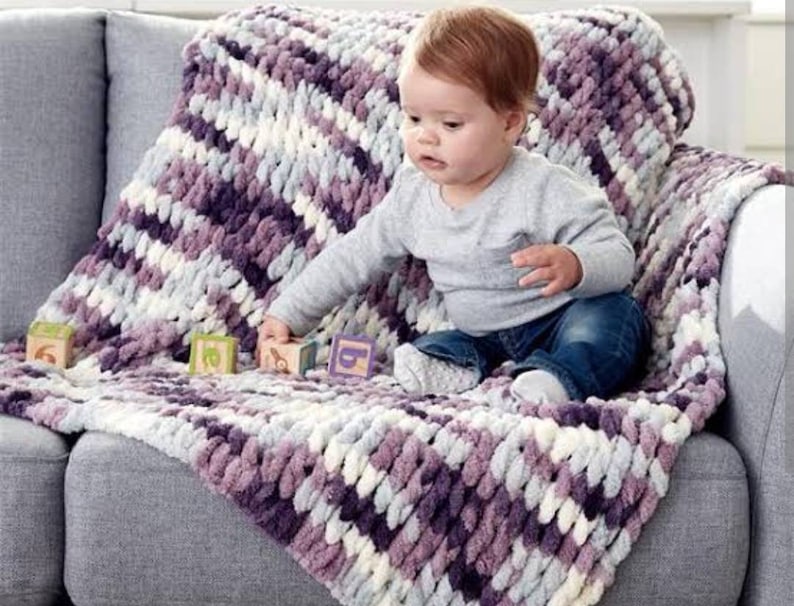 May include: A baby sitting on a couch with a purple and white striped knitted blanket. The blanket is made of chunky yarn and has a soft, fluffy texture.