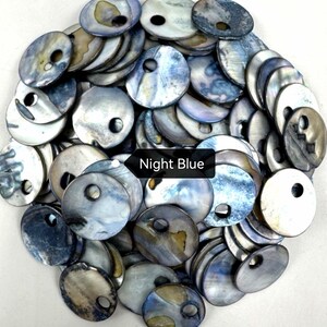 May include: A collection of round, iridescent mother-of-pearl buttons in shades of blue, gray, and silver. Each button features a single off-center hole for attachment. A black rectangular label with the white text Night Blue is visible.