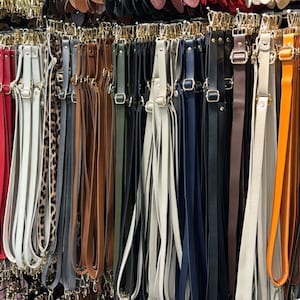 Puede incluir: Una variedad de correas de bolso coloridas cuelgan de un expositor. Las correas están hechas de cuero y otros materiales, en colores como rojo, blanco, marrón, negro y naranja. Algunas correas tienen herrajes dorados.