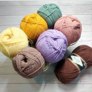 May include: A collection of eight balls of yarn in various colours, including yellow, light yellow, purple, brown, green, and pink. The yarn is arranged in a circle on a white wooden surface. The yarn is labelled "Alize Superlana Maxi"