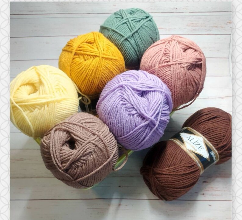 May include: A collection of eight balls of yarn in various colours, including yellow, light yellow, purple, brown, green, and pink. The yarn is arranged in a circle on a white wooden surface. The yarn is labelled "Alize Superlana Maxi"