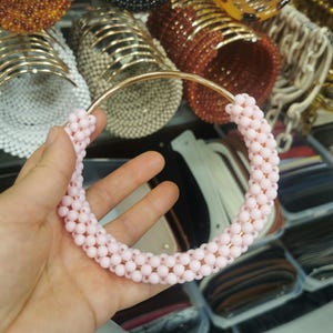 May include: A light pink beaded necklace with a gold-colored metal frame. The necklace is made of small, round beads and is held in a hand. Other beaded bracelets and necklaces in various colors are visible in the background.