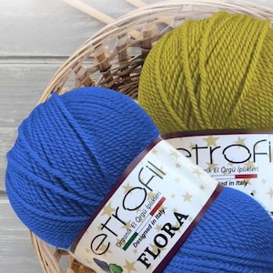 Könnte beinhalten: Zwei Knäuel Garn, eines blau und eines gelb, mit dem Etikett "etrofil FLORA Organic El Örgü İplikleri Designed in Italy" auf dem blauen Knäuel.