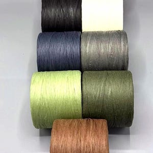 Op de afbeelding: Een verzameling kleurrijke raffia lintrollen in diverse tinten, waaronder geel, zwart, wit, marineblauw, grijs, groen, olijf en bruin. De rollen zijn verticaal gerangschikt, waarbij hun levendige kleuren en texturen worden getoond.
