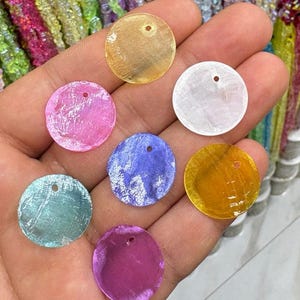 May include: A collection of seven circular, flat shell beads in various translucent colours including yellow, white, pink, blue, purple, and teal. Each bead features a single small hole near the top edge for stringing, with a smooth, iridescent surface.