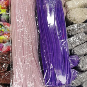 May include: Two bundles of craft supplies are visible, one pink and one purple, both in clear plastic packaging. The pink bundle is on the left, and the purple bundle is on the right. Other craft supplies in various colours are visible in the background.