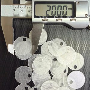 May include: A collection of circular, translucent white acrylic discs, each featuring a single small hole near the edge. A digital caliper is positioned over one disc, displaying a measurement of 20.00 mm on its screen, confirming the diameter.