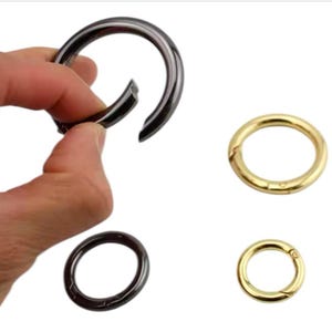 Può includere: Un assortimento di anelli a molla in metallo di vari colori. Una mano apre un grande anello grigio scuro. Sono visibili anche due anelli color oro e un anello grigio scuro, tutti con un design circolare e un cancello a molla.