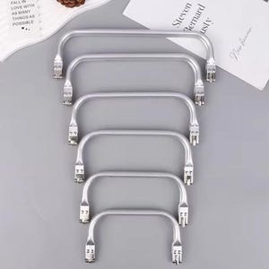 May include: A stack of five silver-colored metal bag handles. Each handle has a rectangular shape with curved sides and a mounting bracket on each end. The handles are arranged vertically against a gray background. Text on paper reads "FALL IN LOVE WITH AS MANY THINGS AS POSSIBLE."