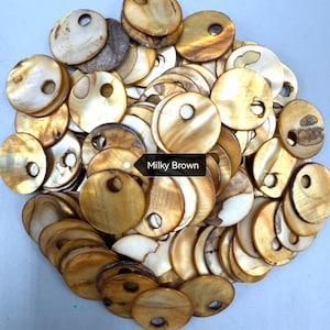 May include: A collection of circular, flat shell buttons in shades of milky brown, tan, and cream. Each button features a smooth, iridescent surface with a single off-center hole for attachment. The buttons are arranged in a dense, overlapping pile.
