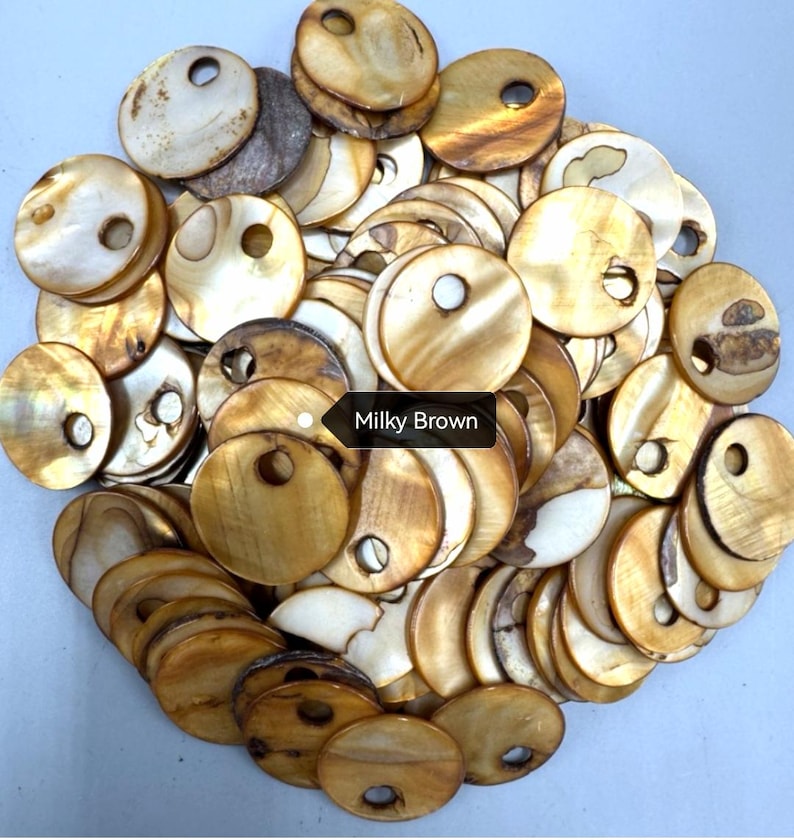 May include: A collection of circular, flat shell buttons in shades of milky brown, tan, and cream. Each button features a smooth, iridescent surface with a single off-center hole for attachment. The buttons are arranged in a dense, overlapping pile.