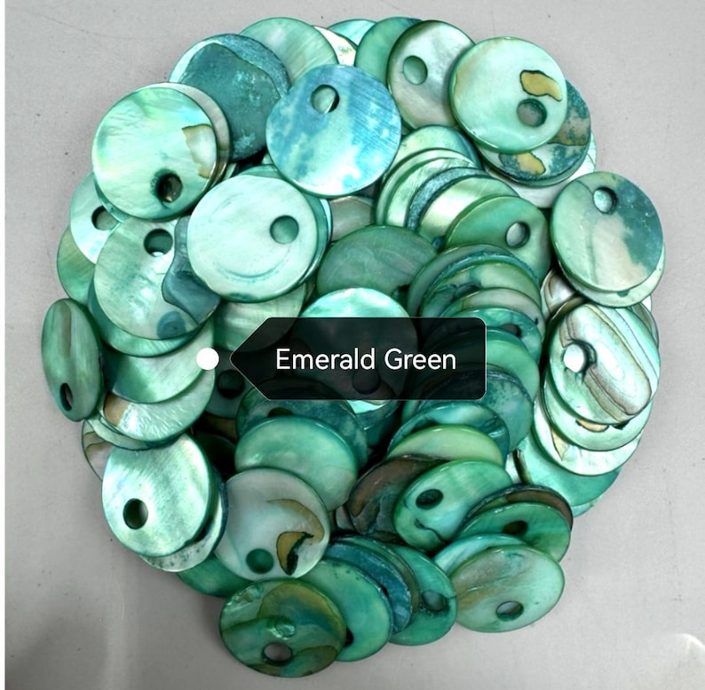 May include: A collection of round, emerald green mother-of-pearl buttons, each featuring a single off-center hole. The buttons display natural iridescent variations and swirling patterns in shades of green, with a black label reading Emerald Green.