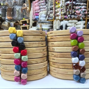 May include: Stacks of wooden embroidery hoops with colorful, geometric beads attached. The hoops are made of light-colored wood and are arranged in neat piles. The beads are in various colors, including yellow, blue, red, and pink. The background shows craft supplies.