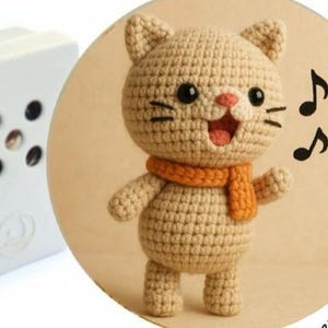 May include: A crocheted cat doll with an orange scarf and musical notes. The beige cat has black eyes, whiskers, and a pink nose and mouth. The doll is standing in front of a tan circle, with two white boxes in the background.