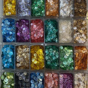 May include: A collection of small, round, iridescent shell buttons organized into twenty-four clear plastic rectangular compartments. The buttons are sorted by color, featuring shades of yellow, blue, green, orange, white, brown, purple, and pink.