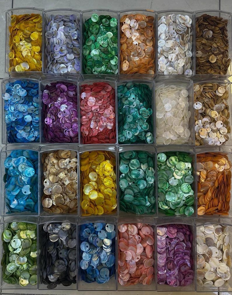 May include: A collection of small, round, iridescent shell buttons organized into twenty-four clear plastic rectangular compartments. The buttons are sorted by color, featuring shades of yellow, blue, green, orange, white, brown, purple, and pink.