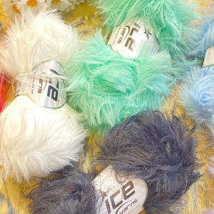 Peut inclure: Plusieurs pelotes de laine duveteuse aux couleurs pastel, dont blanc, vert menthe et gris. Chaque pelote porte une étiquette "ICE YARNS". La laine est disposée sur une surface en bois clair, avec des marguerites artificielles en arrière-plan.