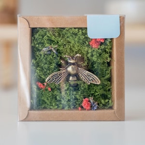 May include: A small, square box with a clear lid, filled with green moss and a gold-colored bee figurine. The box is made of brown cardboard and has a light blue label on the top.