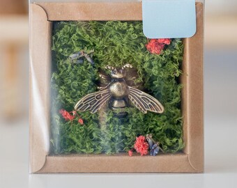 May include: A small, square box with a clear lid, filled with green moss and a gold-colored bee figurine. The box is made of brown cardboard and has a light blue label on the top.