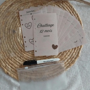 Peut inclure: Ensemble de feuilles de papier roses avec l'inscription "Challenge 12 mois 1200€" et des illustrations de cœurs, accompagnées d'un marqueur noir et blanc et d'une pochette plastique transparente. Le tout est disposé sur un tapis tressé.