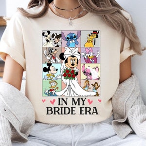 Può includere: Una maglietta bianca con una grafica di personaggi Disney, tra cui Topolino, Minnie Mouse, Stitch e Paperino. Il testo "In My Bride Era" è stampato sotto la grafica.