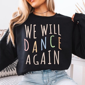 May include: A black sweatshirt with the text "WE WILL DANCE AGAIN" in colourful letters. The letters are in different colours, including pink, blue, green, and yellow.