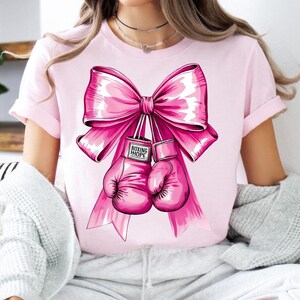 May include: Pink t-shirt with a large pink bow and two pink boxing gloves. The gloves have the words "Boxing Hope" written on them.