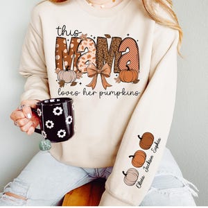 Könnte beinhalten: Beiges Sweatshirt mit dem Text "this MAMA loves her pumpkins" in einem Herbst-Design. Der Ärmel zeigt Kürbisgrafiken und die Namen Olivia Jackson Sophia. Eine schwarze Tasse mit weißen Blumenmustern wird gehalten.