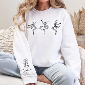 May include: White sweatshirt featuring three ballet dancer illustrations in black. The sleeve displays a ballet shoe graphic and the name "Sophia". The design is simple and elegant, perfect for ballet enthusiasts.