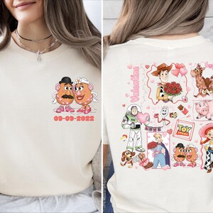 Könnte beinhalten: Cremefarbenes T-Shirt mit einem Cartoon-Design von Mr. und Mrs. Potato Head mit dem Datum 09-03-2022. Auf der Rückseite des Shirts sind verschiedene Toy Story-Charaktere und das Wort "Valentine" in Pink abgebildet.