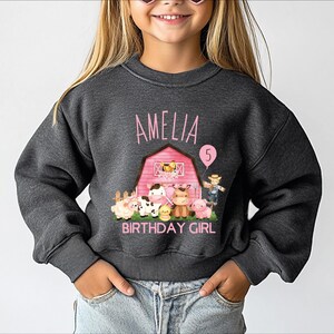 A young girl wearing a gray sweatshirt with a birthday-themed design, including a barn, farm animals, and the text "Amelia 5".