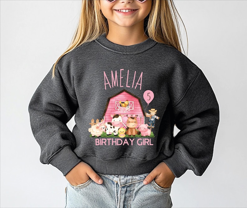 A young girl wearing a gray sweatshirt with a birthday-themed design, including a barn, farm animals, and the text "Amelia 5".