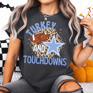 Op de afbeelding: Donkergrijs T-shirt met de tekst "TURKEY AND TOUCHDOWNS" in lichtblauw. Het ontwerp bevat een bruine American football, een lichtblauwe ster en een luipaardprint achtergrond.