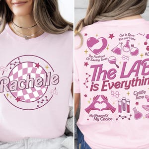 Pode incluir: Camiseta rosa com um design circular xadrez rosa e branco e o nome "Rachelle" em uma fonte retrô. A parte de trás da camiseta tem um design rosa e branco com o texto "The Lab is Everything" e outros gráficos temáticos científicos.